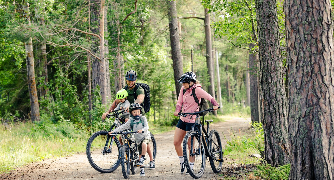 Familj med två vuxna och två barn på cyklar i skogsmiljö